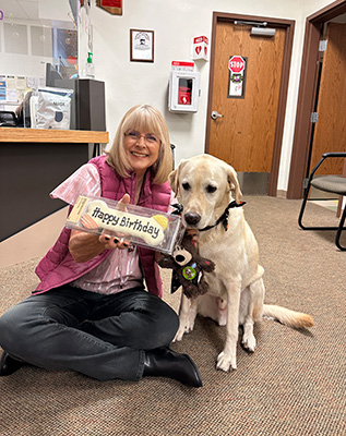 Staff member holding a bone shaped birthday cookie for Darrien the Therapy Dog. Darrien is sitting beside her.