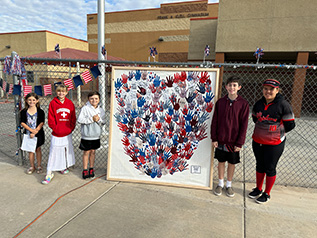 Five students standing beside chainlink fence with large framed heart image made out of handprints